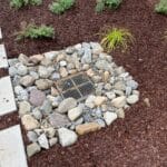 A metal drain grate is surrounded by various sized rocks, set within a landscaped area with mulch, stepping stones, and small green plants.