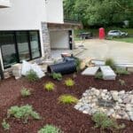 A landscaped yard with mulch, rocks, and small plants beside a modern house; construction materials and a large black pipe are scattered nearby.