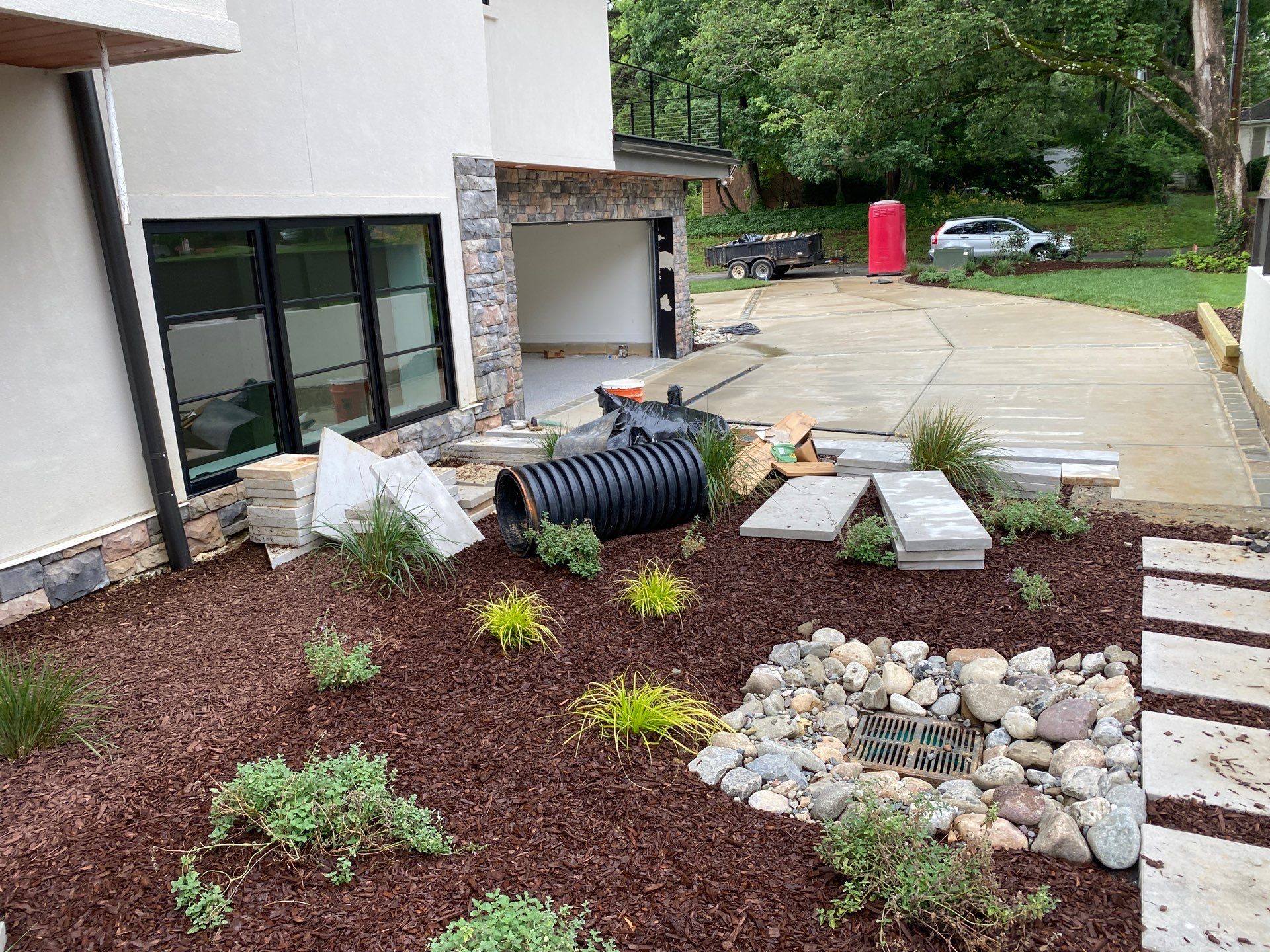A landscaped yard with mulch, rocks, and small plants beside a modern house; construction materials and a large black pipe are scattered nearby.