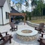 A backyard patio with Adirondack chairs around a circular brick fire pit, a low stone retaining wall, and a pergola with a seating area in the background.