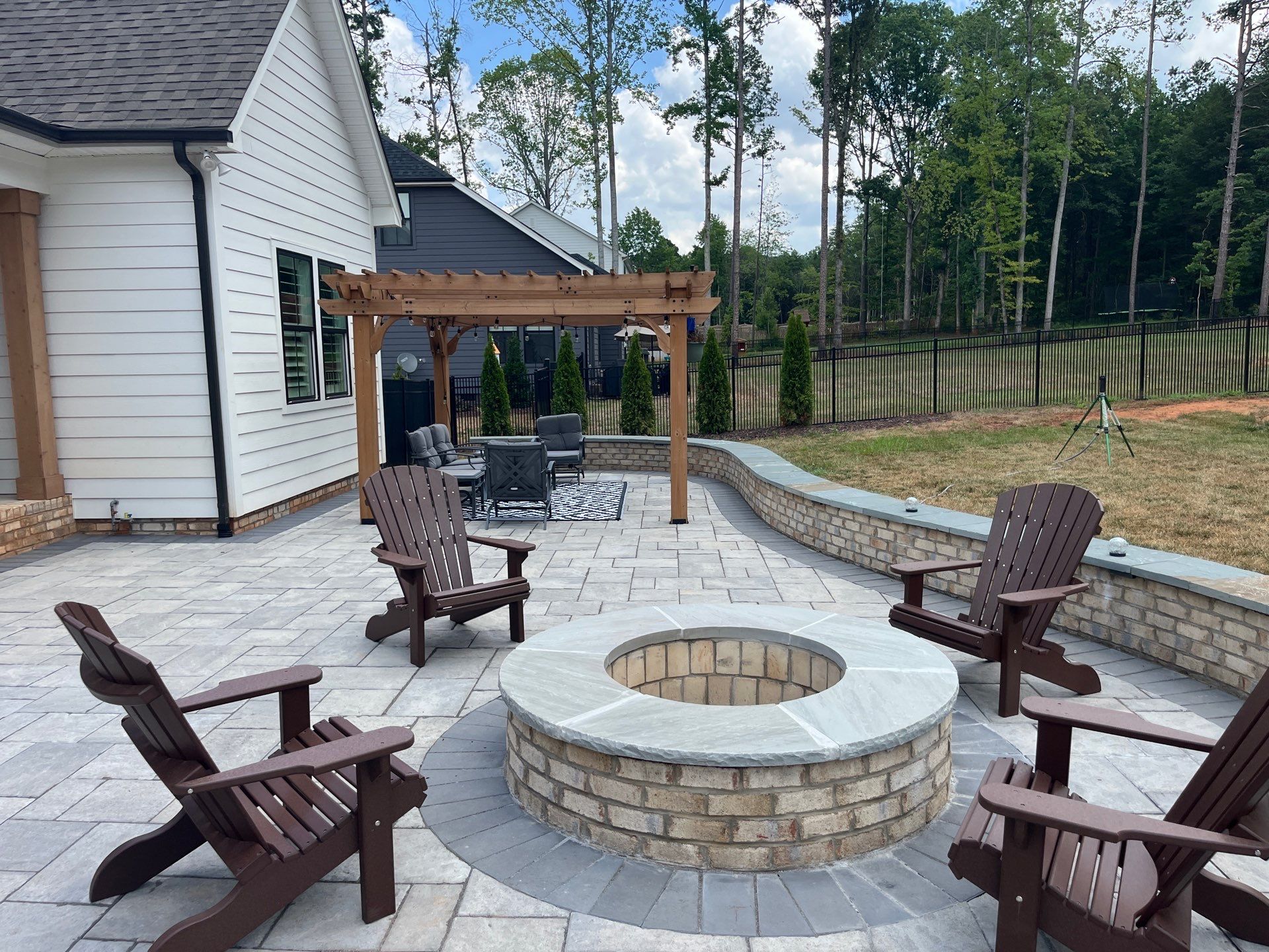 A backyard patio with Adirondack chairs around a circular brick fire pit, a low stone retaining wall, and a pergola with a seating area in the background.