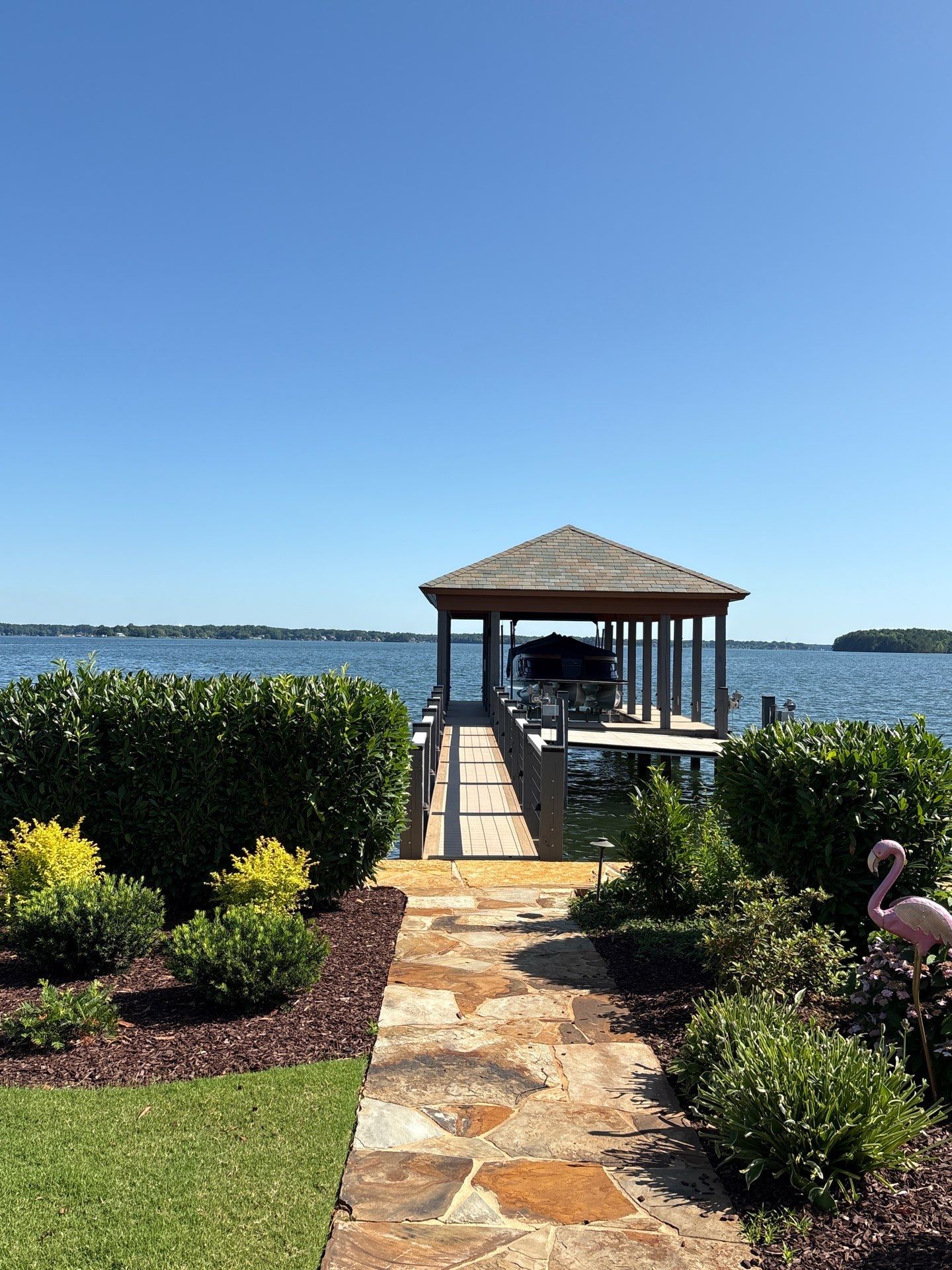 A stone path leads through landscaped bushes to a covered dock with a boat on a lake under a clear blue sky; a pink flamingo lawn ornament is on the right.