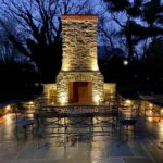 Outdoor stone fireplace with accent lighting, surrounded by metal chairs and tables on a stone patio at dusk.