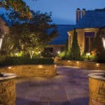 A well-lit stone patio with decorative lamp posts, pathway lights, trees, shrubs, and a large stone house in the background at dusk.