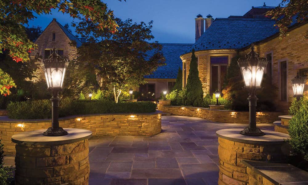 A well-lit stone patio with decorative lamp posts, pathway lights, trees, shrubs, and a large stone house in the background at dusk.