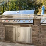 A built-in stainless steel outdoor grill with drawers and a side burner, set in a stone patio countertop, with trees in the background.