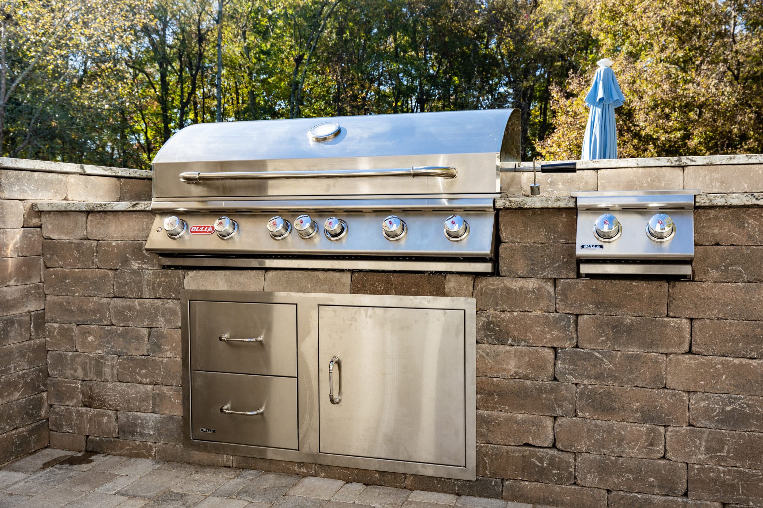 A built-in stainless steel outdoor grill with drawers and a side burner, set in a stone patio countertop, with trees in the background.