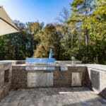 Outdoor kitchen with a stainless steel grill and storage cabinets, surrounded by stone countertops and trees in the background.