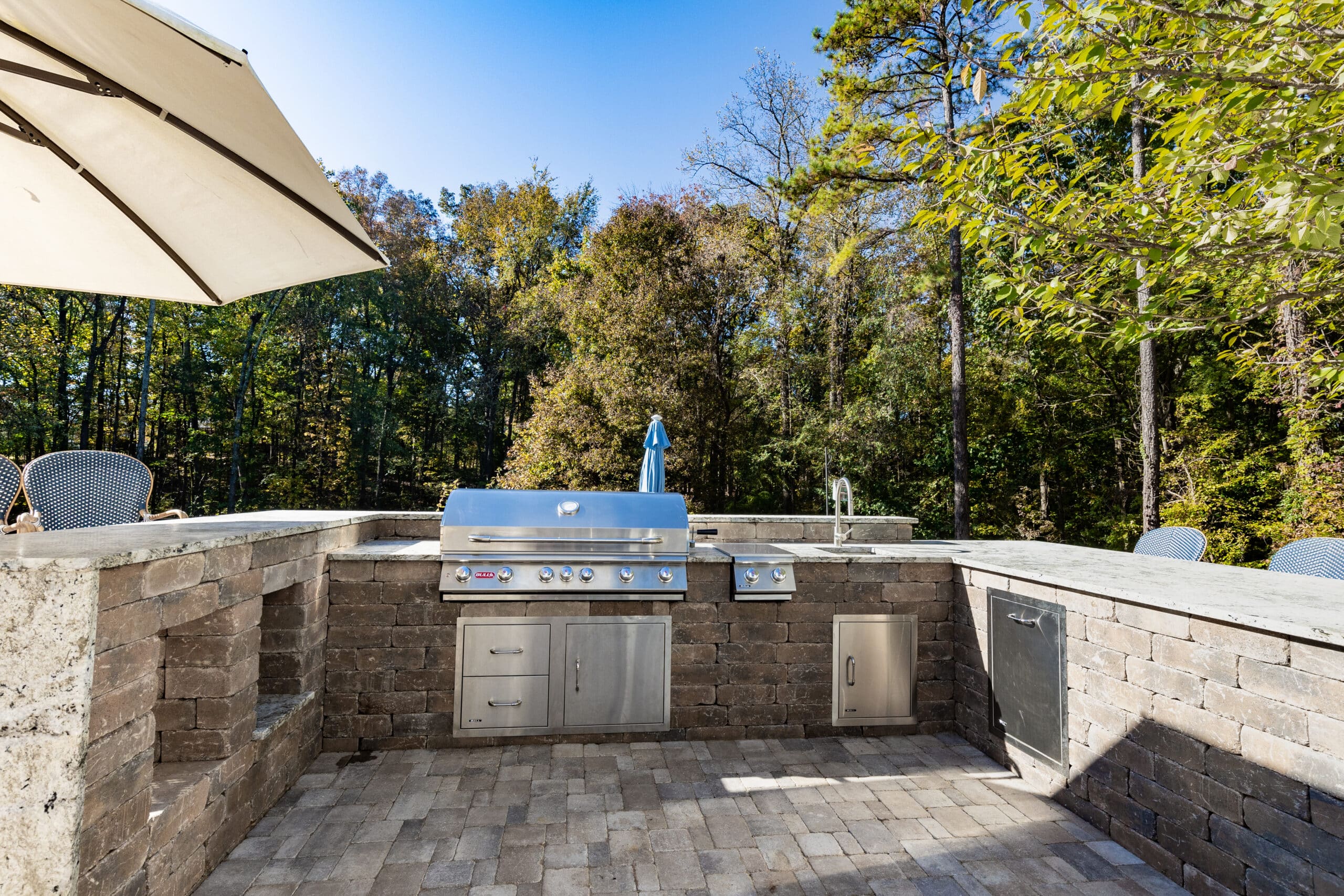 Outdoor kitchen with a stainless steel grill and storage cabinets, surrounded by stone countertops and trees in the background.