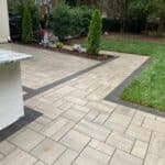 Backyard with a paved patio, bordered by dark bricks, adjacent to a grassy lawn and landscaped flower bed with small trees and purple flowers.