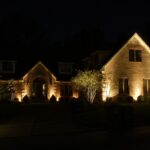 A brick house at night with exterior lights illuminating the front walls, trees, and shrubs. The surroundings are dark except for the lit areas.