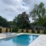 A backyard swimming pool with a built-in hot tub, surrounded by a stone patio, privacy fence, and trees on a cloudy day.