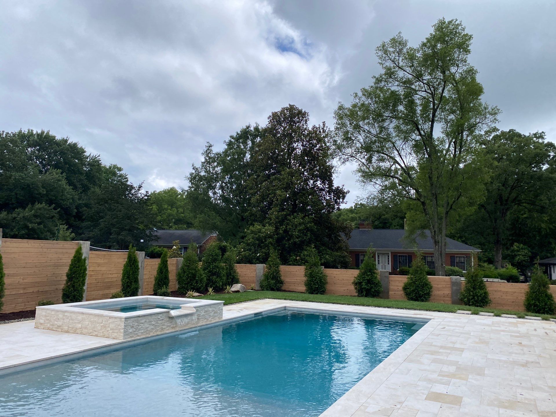 A backyard swimming pool with a built-in hot tub, surrounded by a stone patio, privacy fence, and trees on a cloudy day.