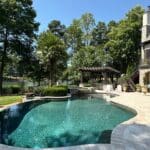 A backyard with a curved swimming pool, stone patio, lounge chairs, a pergola, and trees in the background on a clear, sunny day.