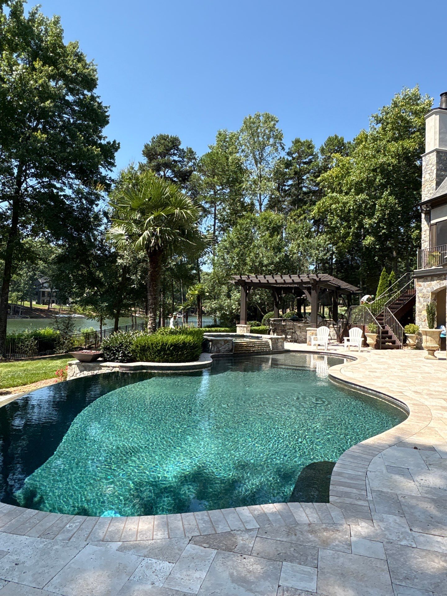 A backyard with a curved swimming pool, stone patio, lounge chairs, a pergola, and trees in the background on a clear, sunny day.