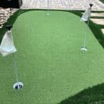 A backyard putting green with three holes marked by white flags, surrounded by artificial grass, stones, and stepping stones.