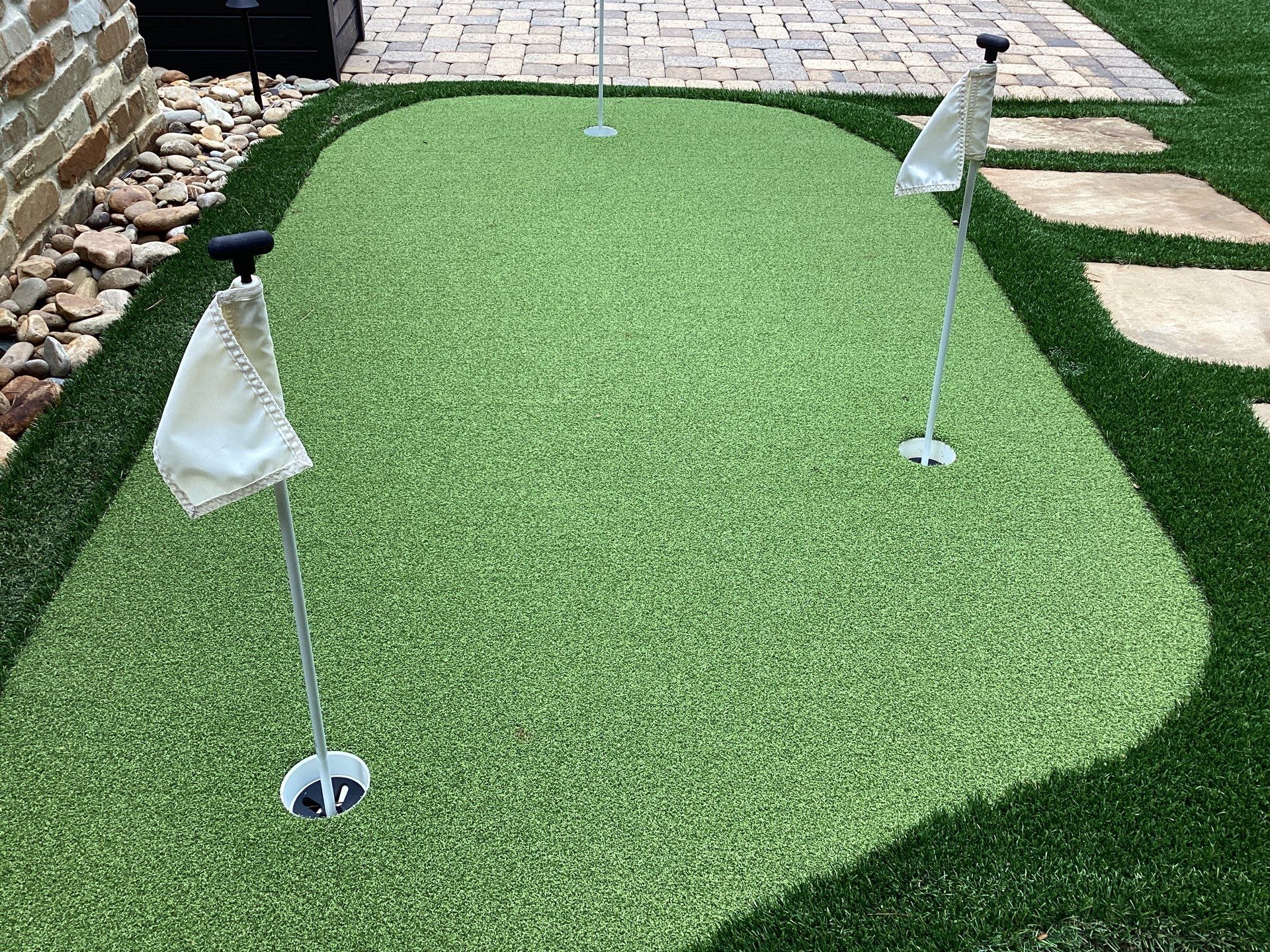 A backyard putting green with three holes marked by white flags, surrounded by artificial grass, stones, and stepping stones.