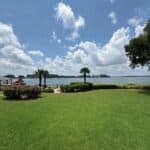A well-kept grassy lawn with hedges and palm trees overlooks a calm lake under a partly cloudy sky. A wooden dock and gazebo are visible on the left side near the water.