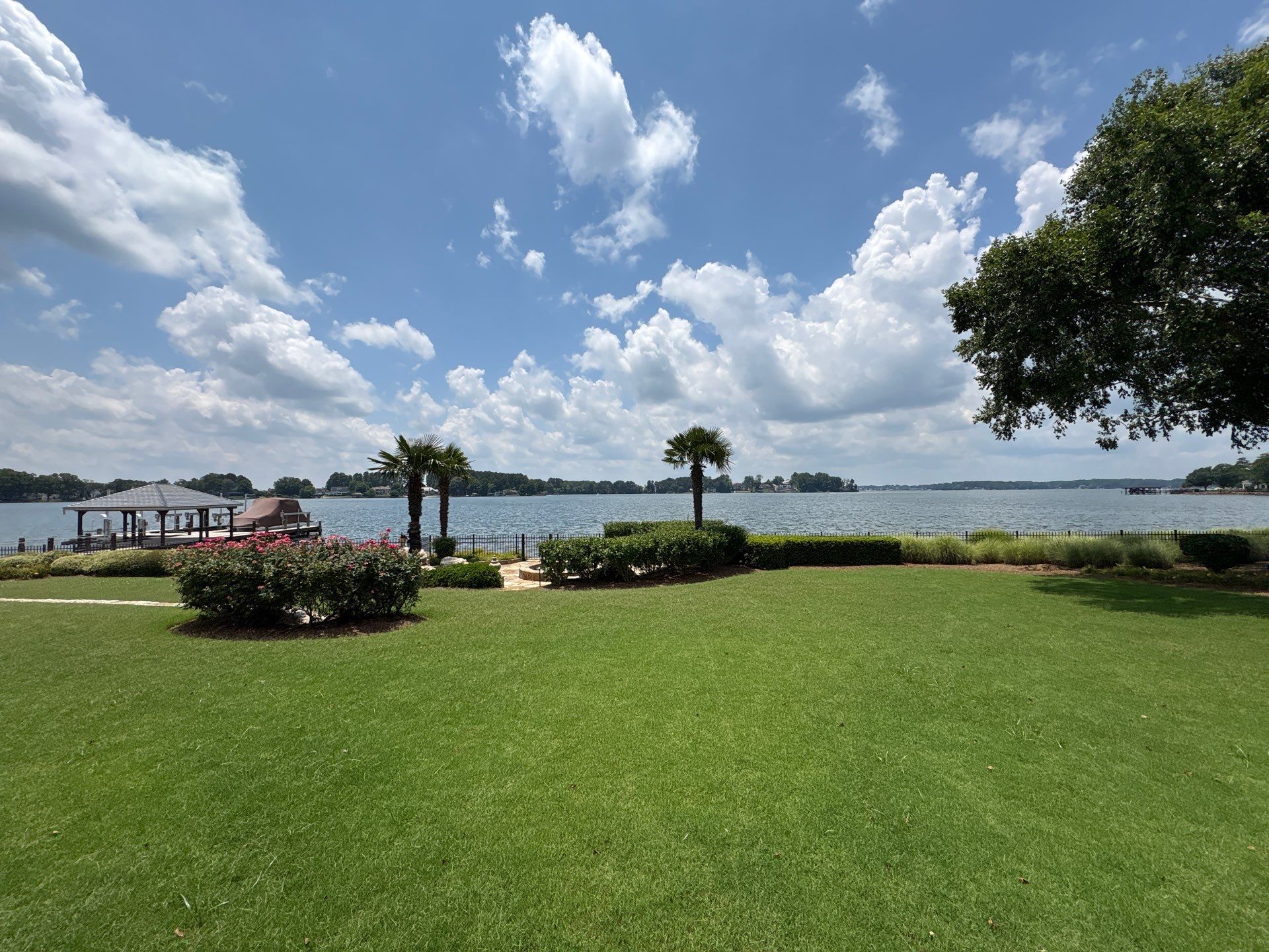 A well-kept grassy lawn with hedges and palm trees overlooks a calm lake under a partly cloudy sky. A wooden dock and gazebo are visible on the left side near the water.