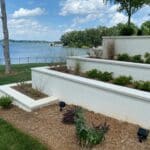 Terraced white planters with shrubs and mulch, overlooking a grassy area and a lake with trees and a partly cloudy sky in the background.
