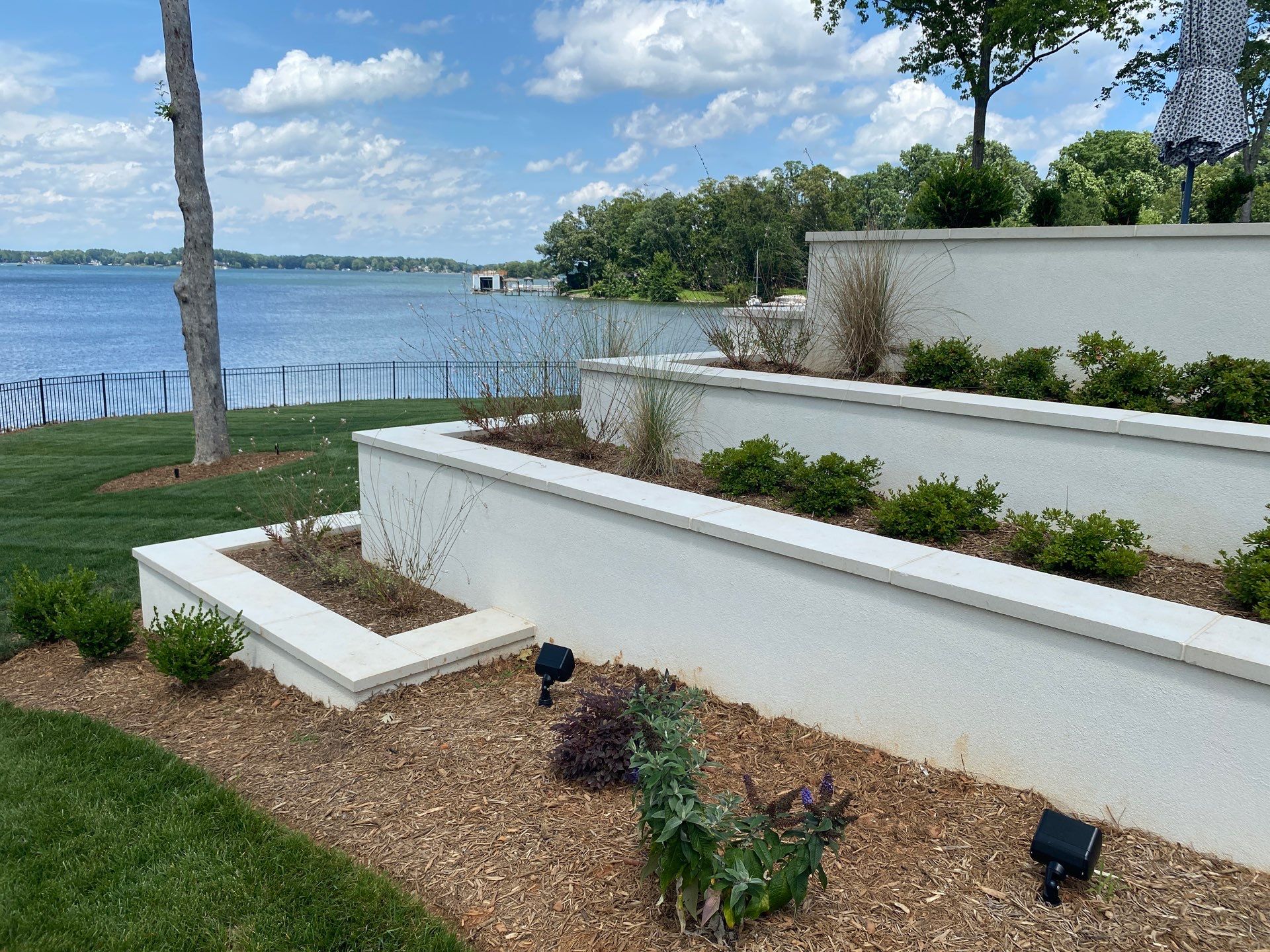 Terraced white planters with shrubs and mulch, overlooking a grassy area and a lake with trees and a partly cloudy sky in the background.