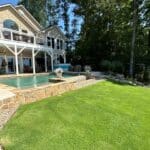 A two-story house with a balcony overlooks a stone-bordered backyard pool, surrounded by a well-maintained lawn and shaded by tall trees.