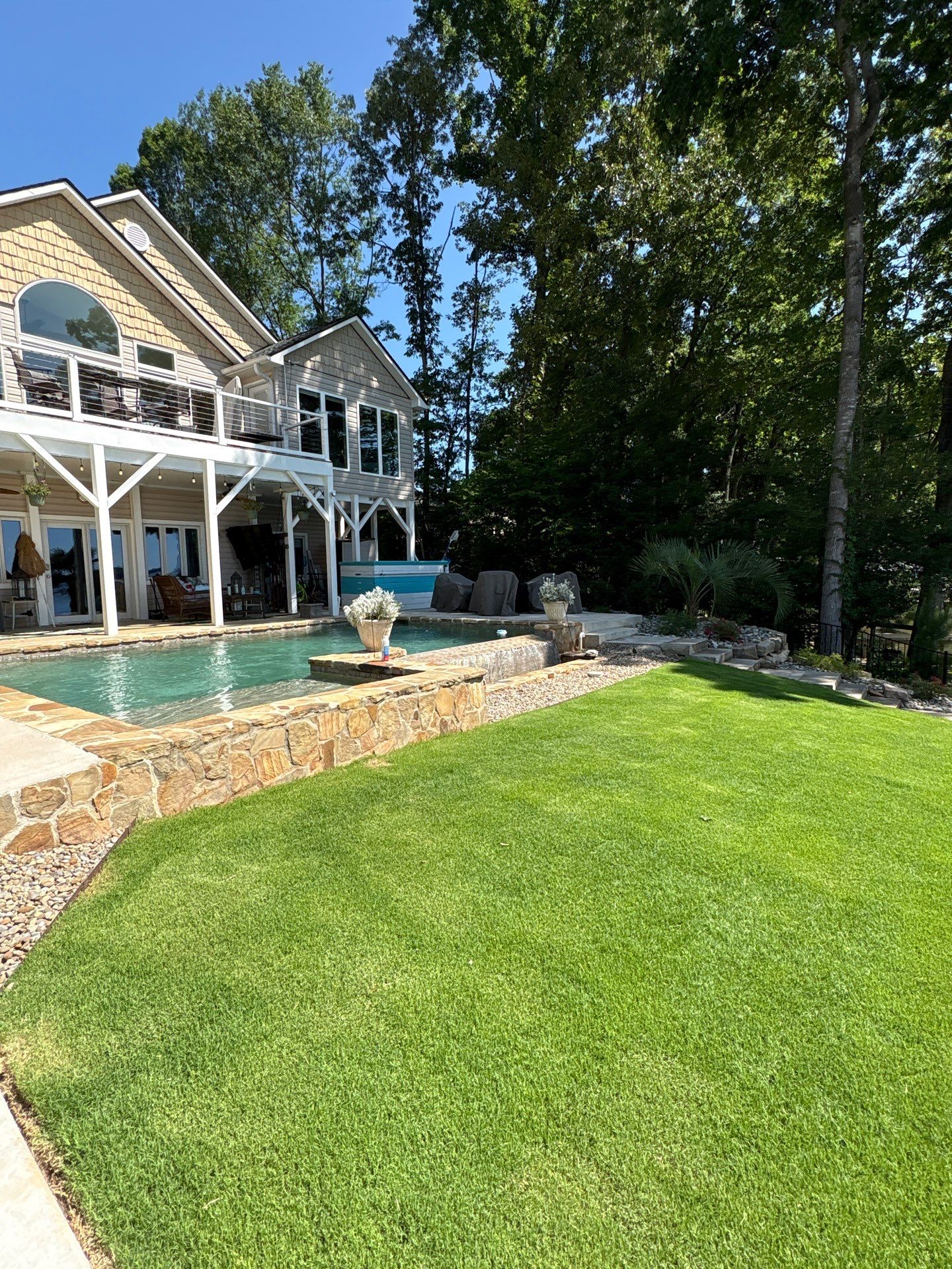 A two-story house with a balcony overlooks a stone-bordered backyard pool, surrounded by a well-maintained lawn and shaded by tall trees.