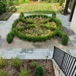 A formal garden with a circular flower bed bordered by trimmed boxwood hedges, surrounded by stone tiles and adjacent to a brick building.