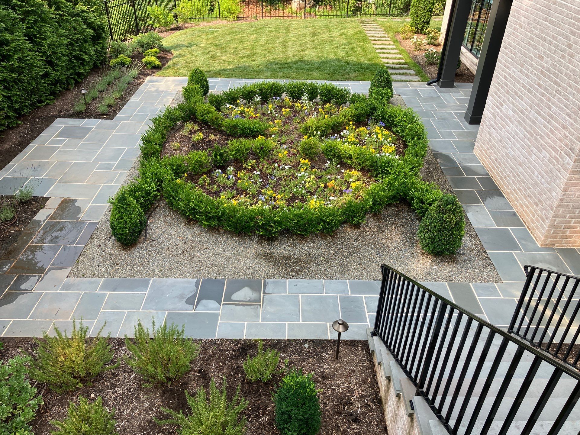 A formal garden with a circular flower bed bordered by trimmed boxwood hedges, surrounded by stone tiles and adjacent to a brick building.