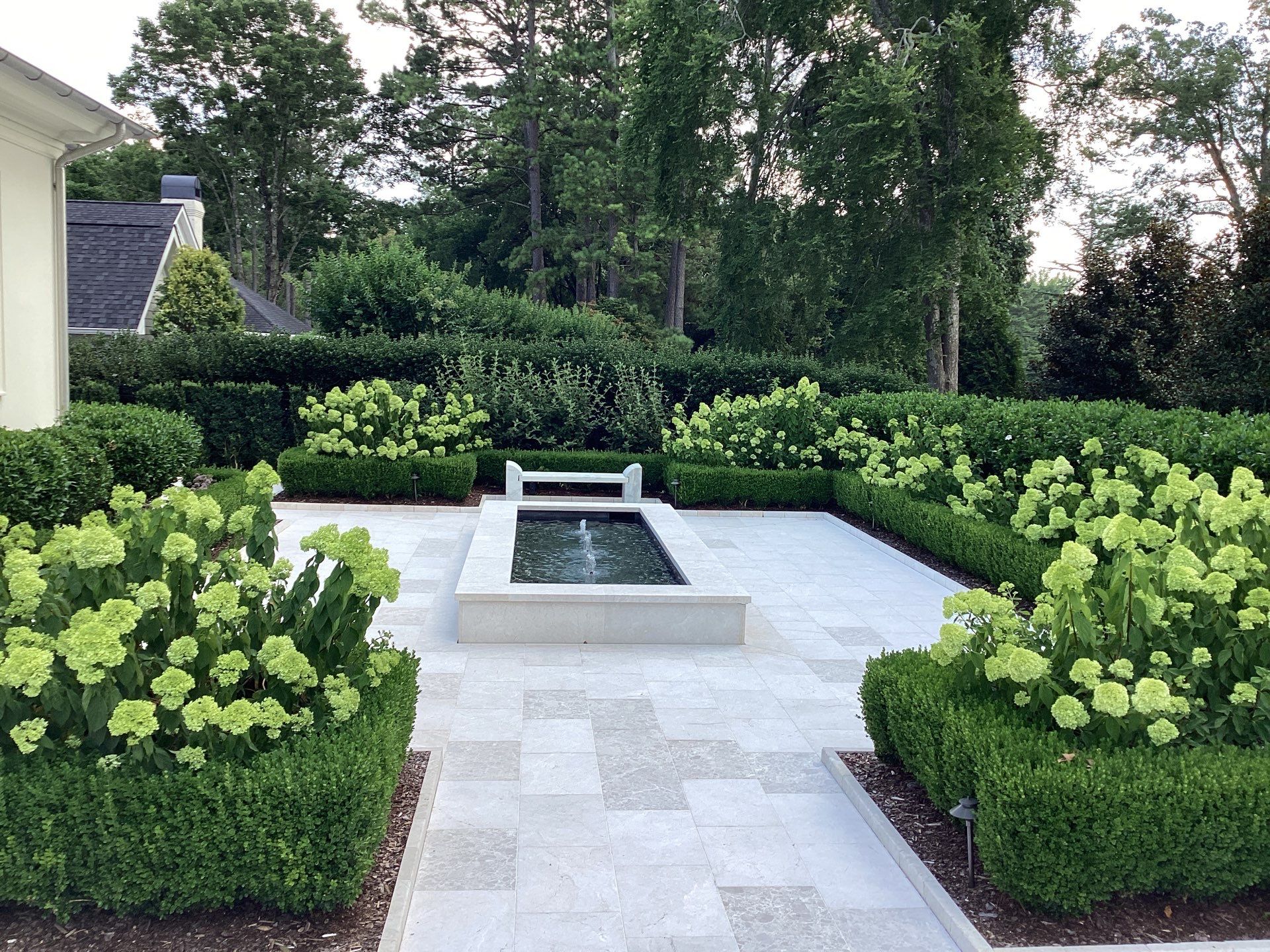 Formal garden with symmetrical boxwood hedges, blooming hydrangeas, tiled pathways, and a rectangular water fountain at the center with a bench beside it. Tall trees in the background.