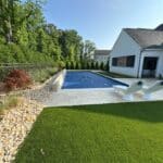 Backyard pool with a blue cover, two white lounge chairs, artificial grass, stone landscaping, and a white house in the background.