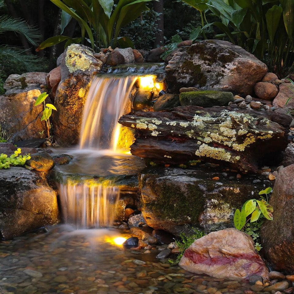 A small waterfall flows over rocks into a pond, illuminated by soft yellow lights, surrounded by green plants and moss.
