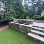 Stone retaining wall and steps beside artificial grass lawn, with a large succulent plant in a raised planter; trees and fence in the background.