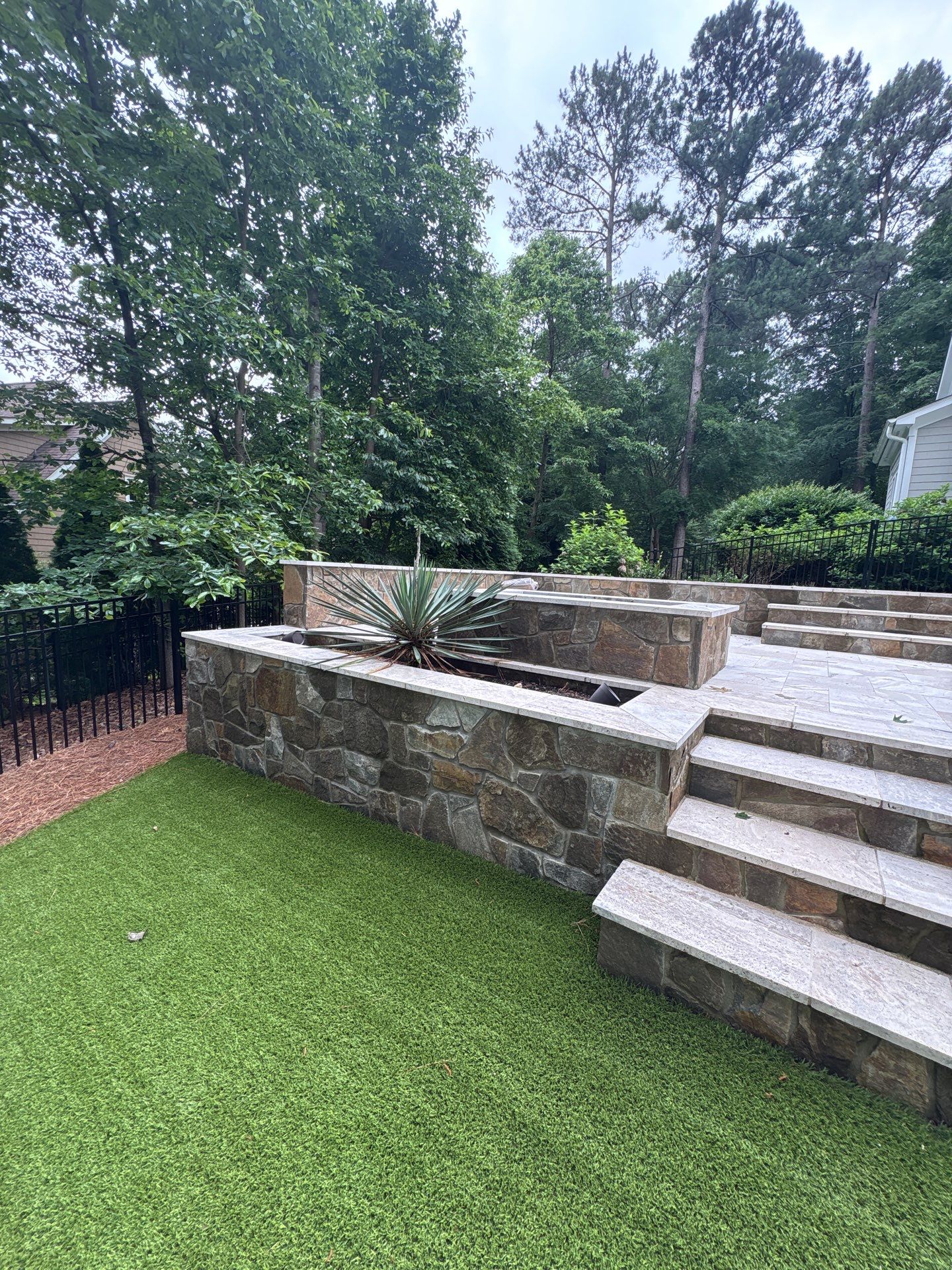 Stone retaining wall and steps beside artificial grass lawn, with a large succulent plant in a raised planter; trees and fence in the background.