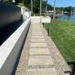 Rectangular stone pavers form a pathway through gravel beside a pool; grass lawn and lake are visible in the background.