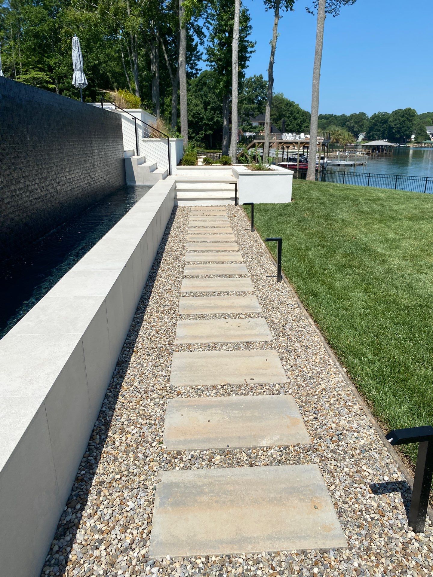 Rectangular stone pavers form a pathway through gravel beside a pool; grass lawn and lake are visible in the background.