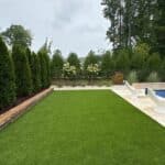 A backyard with artificial turf, lined with tall evergreen trees on the left, white poolside chairs on the right, and a section of a swimming pool visible.