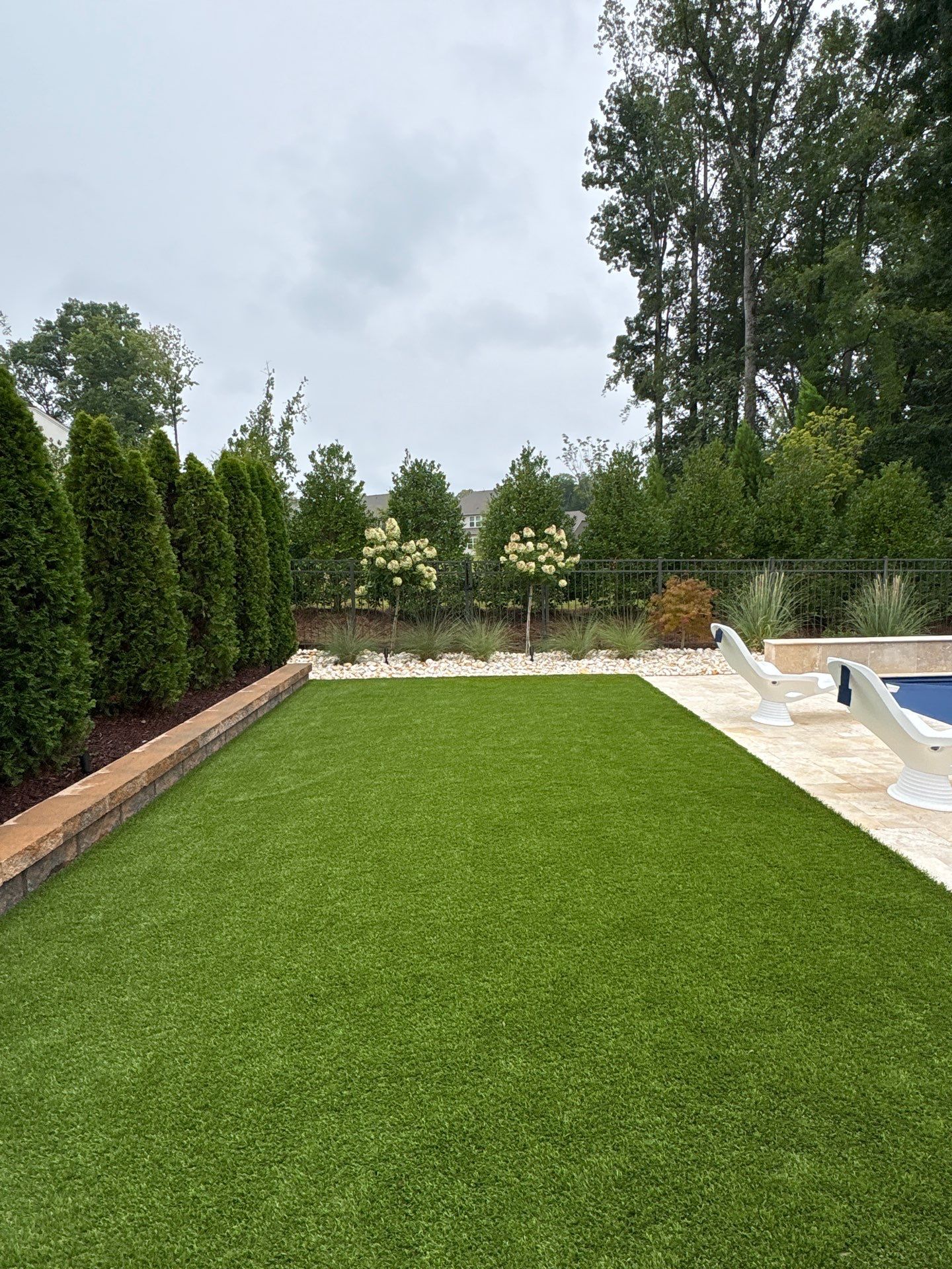 A backyard with artificial turf, lined with tall evergreen trees on the left, white poolside chairs on the right, and a section of a swimming pool visible.