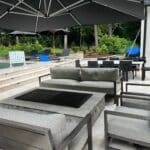 Outdoor patio with gray cushioned seating around a fire pit table, pool loungers, a Connect Four game, and large umbrellas near a swimming pool and greenery.