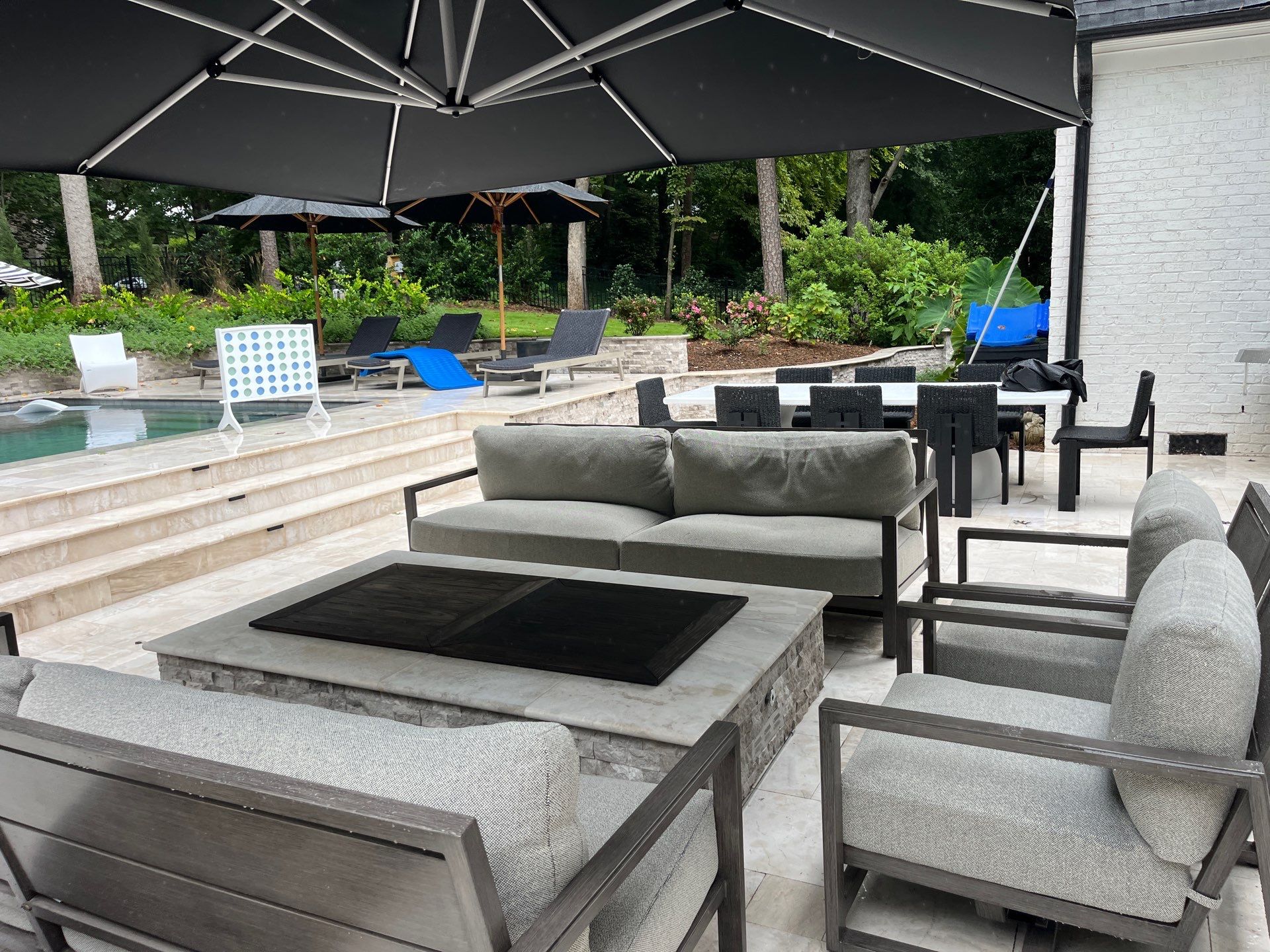 Outdoor patio with gray cushioned seating around a fire pit table, pool loungers, a Connect Four game, and large umbrellas near a swimming pool and greenery.