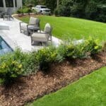 A landscaped backyard with a pool, patio furniture, and a mulched garden bed with green shrubs, bordered by a well-maintained lawn.