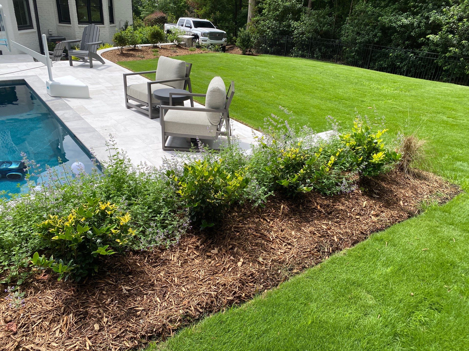 A landscaped backyard with a pool, patio furniture, and a mulched garden bed with green shrubs, bordered by a well-maintained lawn.