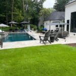 A modern backyard with a rectangular pool, lounge chairs, umbrellas, and patio furniture, surrounded by grass and trees near a white house.