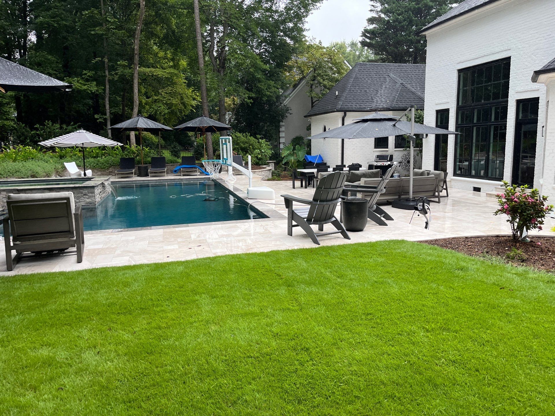 A modern backyard with a rectangular pool, lounge chairs, umbrellas, and patio furniture, surrounded by grass and trees near a white house.