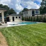 A backyard with a swimming pool, hot tub, lounge chairs, green lawn, and privacy fence, surrounded by modern houses under a partly cloudy sky.