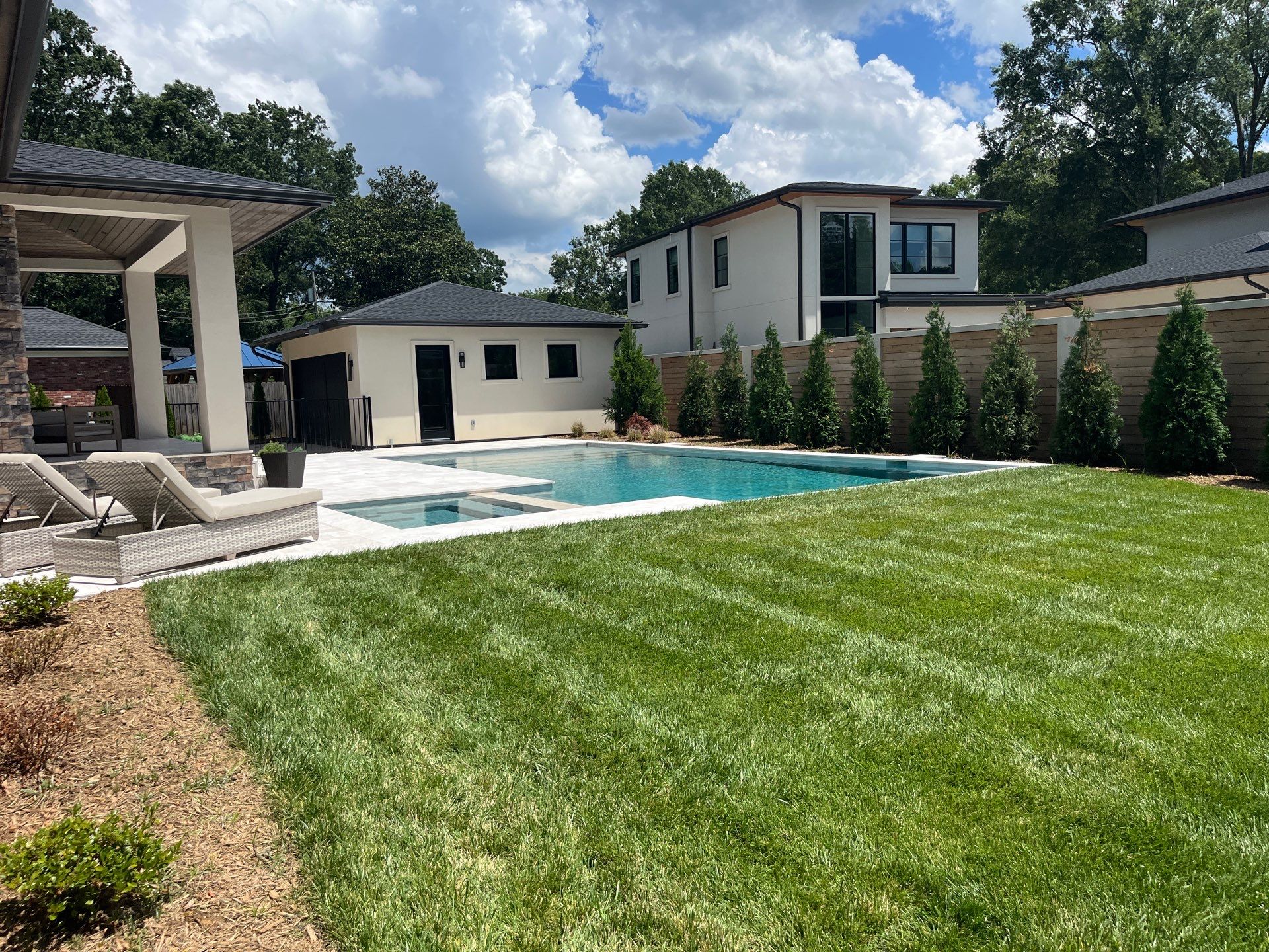 A backyard with a swimming pool, hot tub, lounge chairs, green lawn, and privacy fence, surrounded by modern houses under a partly cloudy sky.
