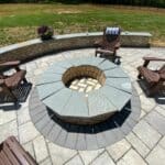 Circular stone fire pit with a surrounding seating wall, four brown Adirondack chairs, and a paver patio. A flower pot sits on the wall, and a grassy yard is in the background.