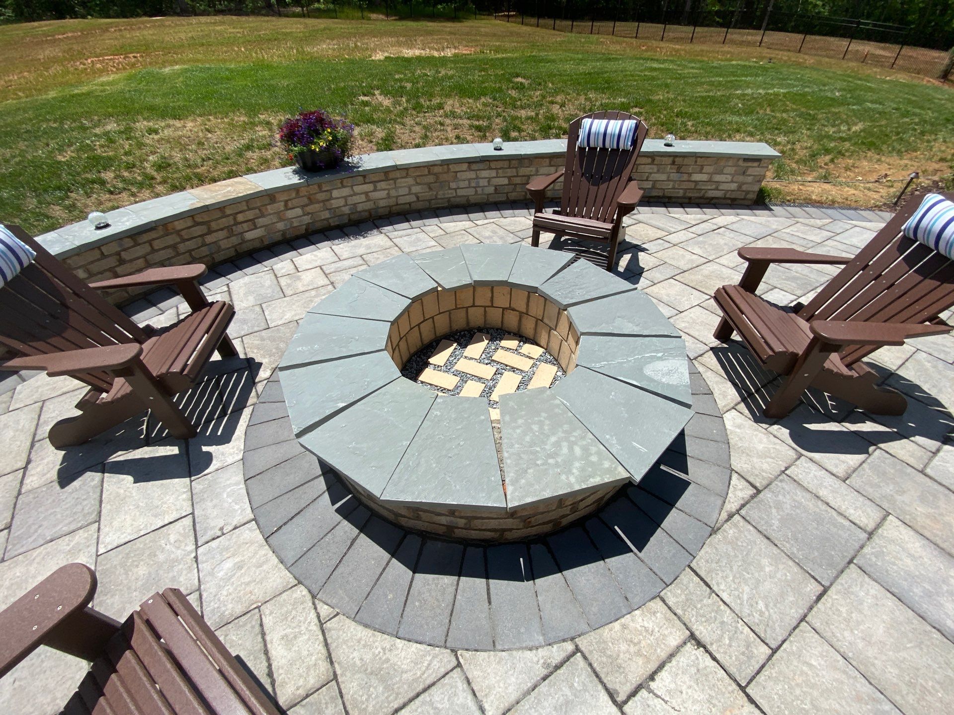 Circular stone fire pit with a surrounding seating wall, four brown Adirondack chairs, and a paver patio. A flower pot sits on the wall, and a grassy yard is in the background.