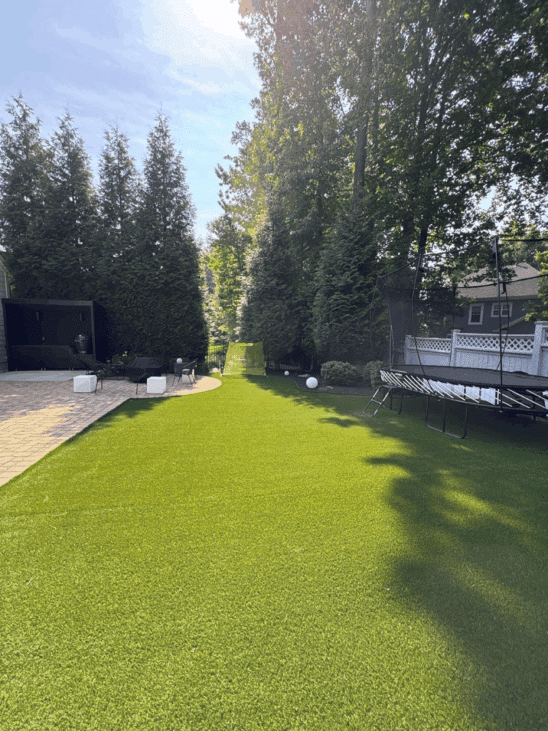 A backyard with artificial grass, a patio area with furniture on the left, a trampoline on the right, and a netted sports goal at the end, surrounded by tall trees and fencing.
