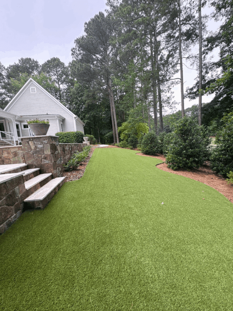 A sloped backyard with artificial turf, bordered by stone steps, mulched plant beds, shrubs, and tall pine trees near a white house.
